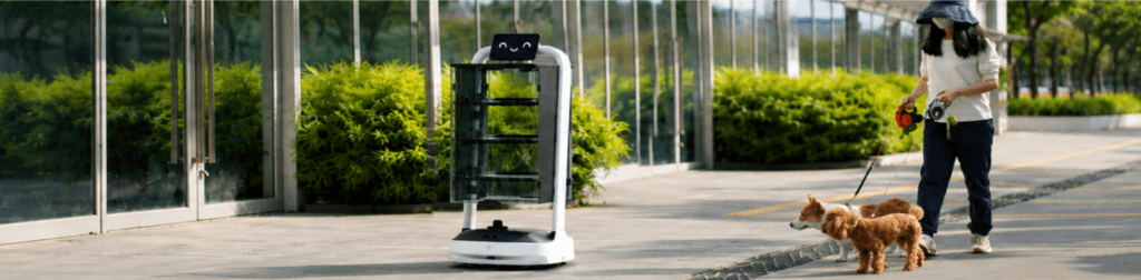 Kiwi autonomous delivery robot operating outdoors along a modern office-campus walkway, with a pedestrian walking two dogs nearby. The robot’s transparent display cabinet is showcased in a real-world service environment, surrounded by glass architecture, landscaped greenery, and natural daylight.