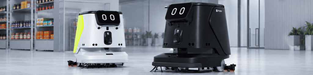 Two autonomous SHOP CC1 commercial cleaning robots operating in a modern industrial facility. A compact white robot and a larger black robot are positioned on a polished warehouse floor, surrounded by storage shelving and glass-walled architecture.